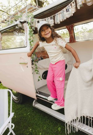 Roze cargobroek, crèmekleurige top met korte mouwen en kleine kleurrijke stippen, krullend haar, staand in een vintage busje met groene planten en feeërieke verlichting.