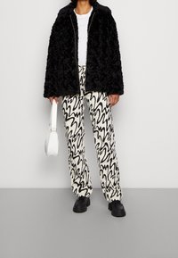 Svart fuskpäls zip-up jacka, vit t-shirt, mönstrade vida byxor i krämfärg med svarta vågiga linjer, svarta skor och vit handväska.