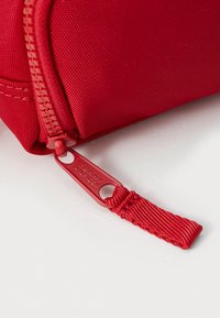 Röd dragkedjepåse med texturerad tyg, som har en märkesdragkedja. Designen inkluderar ett robust nylonnacke för enkel hantering.