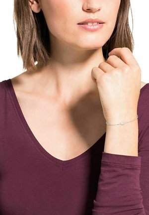 Bracciale in argento con un accento ovale, indossato al polso. Il modello ha capelli castano chiaro e lisci e indossa un top di un profondo colore bordeaux.