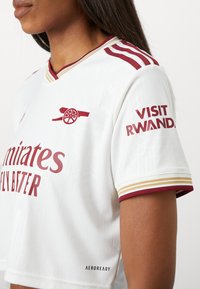 Ženska v belem nogometnem dresu Arsenal z maroon topovskim logotipom, napisom "Emirates Fly Better" in sponzorskimi napisi "Visit Rwanda" na rokavu.