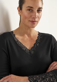 Svart plagg med långa ärmar, med en spets V-ringning och spetsdetaljer vid ärmkanterna, tillverkat av ett mjukt, stretchigt material.