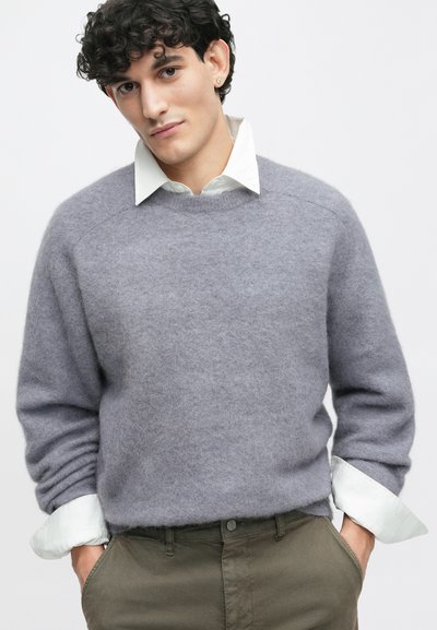 Γκρι μάλλινο πουλόβερ με στρογγυλή λαιμόκοψη, μακριά μανίκια και μαλακή υφή, επάνω από ένα λευκό πουκάμισο με γιακά; ορατά ελαιόχρωμα παντελόνια.