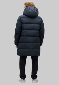 Abrigo de plumas azul marino con capucha desmontable, diseño acolchado y corte holgado. Lleva puesto sobre pantalones negros y zapatillas blancas.