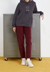 Felpa grigia con cappuccio e design floreale, abbinata a pantaloni in velluto a coste bordeaux e sneaker bianche, in piedi accanto a un armadietto grigio.