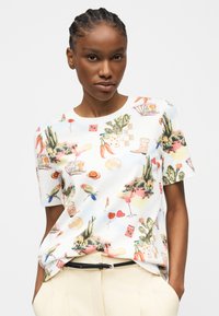 Γυναίκα φοράει λευκό κοντομάνικο πουκάμισο με χρωματιστά σχέδια από κάκτους, πουλιά, καυτερές πιπεριές, καρδιές και κοκτέιλ, συνδυασμένο με μπεζ παντελόνια.