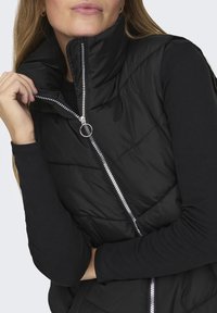 Gilet matelassé noir avec col montant, partiellement ouvert, doté d'une fermeture éclair argentée et d'un tirette circulaire. Haut à manches longues noir en dessous.