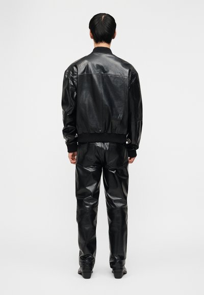 Άνδρας που στέκεται με την πλάτη, φοράει μαύρο δερμάτινο μπουφάν και ταιριαστό δερμάτινο παντ ελόνι, σε απλό φωτεινό φόντο.
