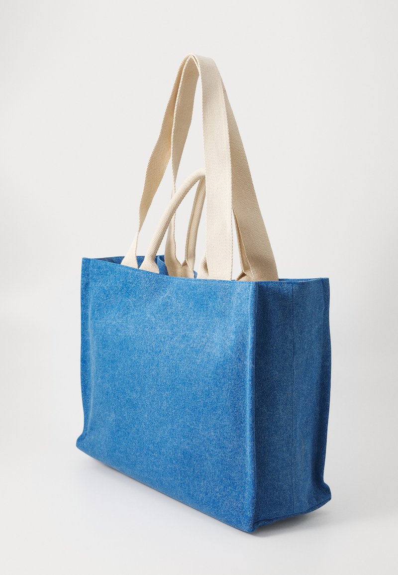 Sac fourre-tout en tissu bleu avec de larges poignées beiges et des sangles d'épaule beiges plus étroites, se tenant debout sur une surface blanche.