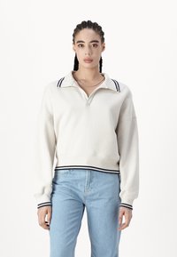 Hanorac alb cu guler, având dungi bleumarin pe guler și mansete. Croi oversized dintr-un material moale și texturat. Asortat cu blugi albaștri deschisi.