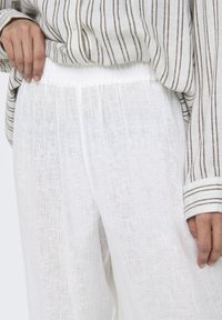 Pantalons en lin blancs et légers avec une ceinture élastique, présentant une finition douce et légèrement texturée. Associés à une chemise à rayures à manches longues.