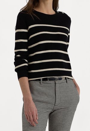 Kvinde iført sort langærmet sweater med hvide vandrette striber og sort-hvide hundetandsbukser med sort bælte, hænderne i lommerne.