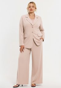 Beige op maat gemaakt pak met een eenrijzige blazer met twee knopen en klepzakken, gecombineerd met broek met wijde pijpen. Soepele textuur.