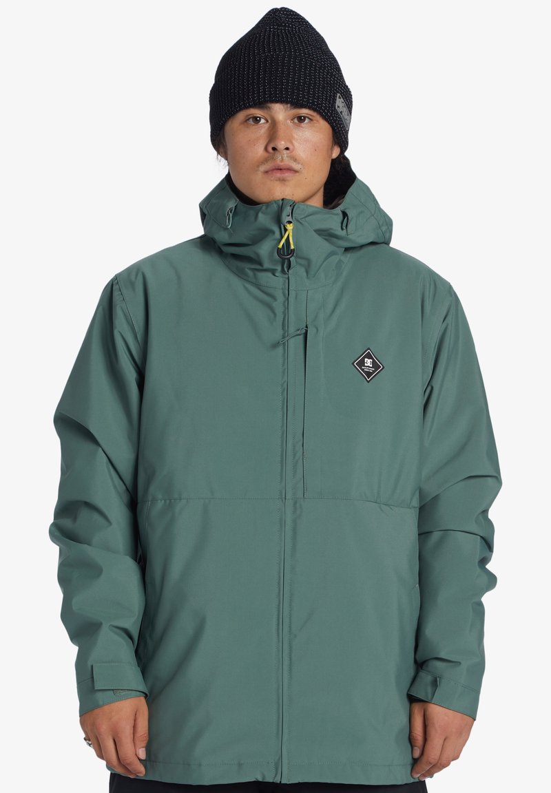 Zelená nepremokavá bunda s voľným strihom, nastaviteľnou kapucňou a predným zipsom. Má logo nášivku na hrudi a dlhé rukávy.