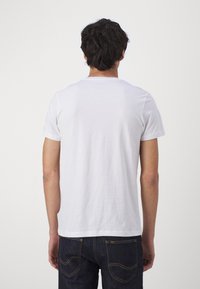 Man met donker haar, die een eenvoudig wit T-shirt met korte mouwen en donkerblauwe spijkerbroek draagt, met zijn gezicht van ons af tegen een effen lichte achtergrond.