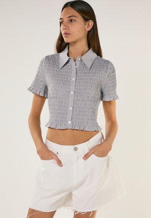 Jonge vrouw met lang bruin haar, gekleed in een grijs gekleurde, kortärmelig, gerimpelde cropped blouse en witte, hooggetailleerde, afgescheurde shorts, handen in de zakken.