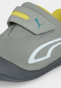 Κοντινή λήψη ενός γκρι sneaker με υφή, κίτρινη επένδυση, λουράκι Velcro, μπλε λογότυπο Puma και λευκό σχέδιο στο πλάι.