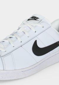 Λευκό αθλητικό Nike με μαύρο λογότυπο swoosh, λευκά κορδόνια και υφασμένο πέλμα, απεικονίζεται σε κοντινή γωνία.