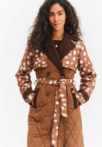 Brązowy pikowany płaszcz typu trencz z brązową, cętkowaną tkaniną na górze i szerokim kołnierzem, z przednim paskiem i dużymi guzikami.