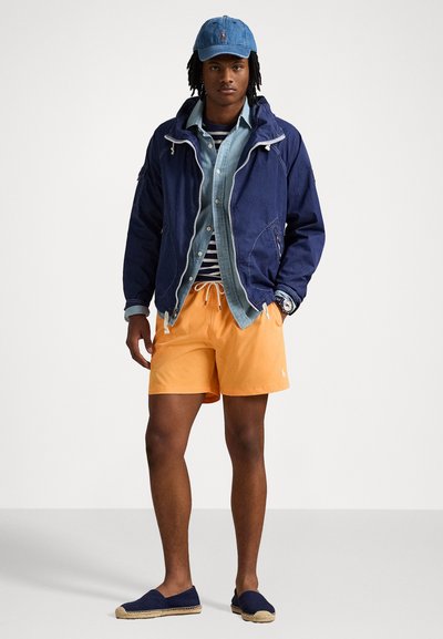 Cortavientos navy con cremalleras blancas, camisa de mezclilla clara, camiseta a rayas por debajo, pantalones cortos naranjas y espadrilles navy con suelas de yute.