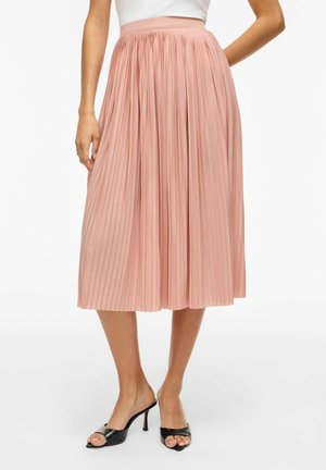 Žena má na sebe svetloružovú plisovanú midi sukňu v kombinácii s čiernymi otvorenými sandálmi na vysokom opätku, stojacu pred bielym pozadím.