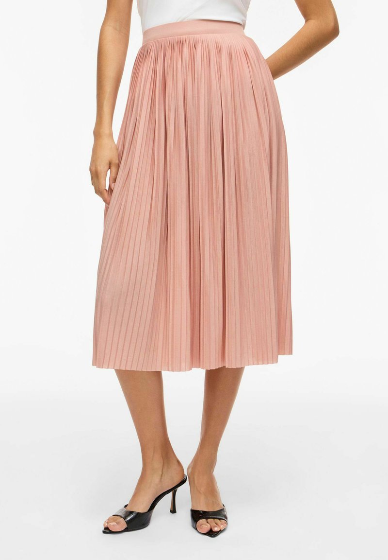 Γυναίκα που φοράει μια ανοιχτό ροζ πλισέ μίντι φούστα σε συνδυασμό με μαύρα ανοιχτά ψηλοτάκουνα σανδάλια, στέκεται μπροστά σε λευκό φόντο.