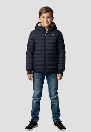 Mørkeblå polstret jakke med hætte, lynlås foran og pelsforet krave, kombineret med blå jeans og sorte sneakers.