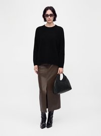 Femme portant un pull noir, une jupe midi en cuir marron avec une fente devant, des bottes noires, des lunettes de soleil foncées, tenant un sac à main noir sur un fond uni.