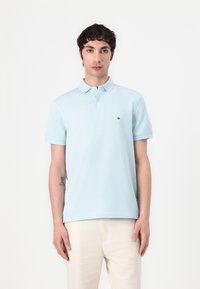 Polo de color azul claro hecho de algodón, con una tapeta de tres botones y un pequeño logo en el pecho. Combinado con pantalones beige claros.