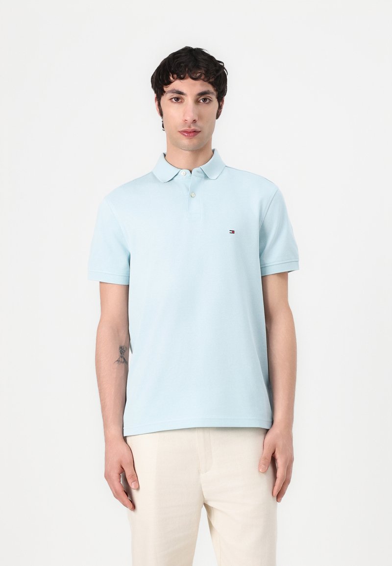 Polo de color azul claro hecho de algodón, con una tapeta de tres botones y un pequeño logo en el pecho. Combinado con pantalones beige claros.