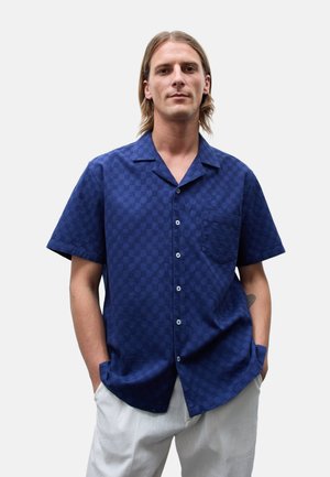 Homme aux cheveux mi-longs portant une chemise bleu foncé à manches courtes boutonnée et un pantalon clair, debout les mains dans les poches.