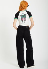 Kurzärmeliges T-Shirt in Weiß und Schwarz mit grünen Akzenten, das auf dem Rücken mit einem bunten Schmetterlingsgrafik verziert ist, kombiniert mit schwarzen weiten Hosen.