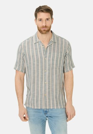 Mand med kort brunt hår og skæg iført en kortærmet skjorte med lodrette striber og lyseblå jeans, stående foran en hvid baggrund.