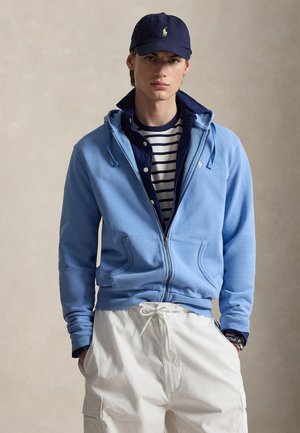 Tânăr purtând șapcă bleumarin, jachetă albastră cu glugă și fermoar suprapusă și cămașă peste tricou în dungi, pantaloni albi cu șnur și mâinile în buzunare.