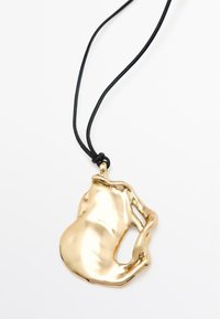 Gouden hanger ketting met een organische, vloeiende vorm, gekenmerkt door een gladde, glanzende oppervlakte, bevestigd aan een zwarte koord met een knoop.