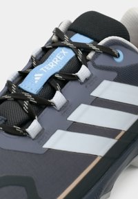 Γκρι αθλητικό παπούτσι με μπλε λεπτομέρειες, διαθέτει υφασμένο επάνω μέρος, τρεις λευκές ρίγες και αξιόπιστο σύστημα κορδονιών για ασφαλή εφαρμογή.