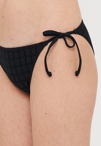 Bas de bikini noir avec un motif texturé, doté de brides à nouer et d'une taille basse. Le matériau est lisse avec un léger élasticité.