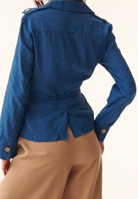 Blå linnekavaj med skräddarsydd passform, med krage, midjebälte och knappaccentsdetaljer på ärmarna. Enfärgad bakdesign.