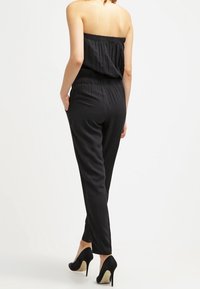 Salopette noire sans bretelles avec une taille élastique et des jambes fuselées, fabriquée dans un tissu lisse. Présente des chaussures noires à talons hauts pour gagner en hauteur.