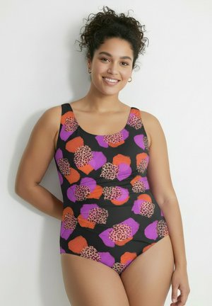 Frau mit lockigem Haar lächelt und trägt einen Badeanzug mit orangefarbenen, violetten und Leopardenmuster-Patches auf schwarzem Hintergrund.