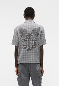 Πουκάμισο με κοντό μανίκι, καρό σε γκρι απόχρωση με φτερό και μπαρόκ σχέδιο μαύρο στο πίσω μέρος. Σχεδίαση με γιακά και άνετη εφαρμογή.