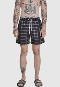 Zwarte zwembroek met een wit rasterpatroon, een elastische tailleband met trekkoord en een ontspannen pasvorm. Gecombineerd met zwarte sandalen.