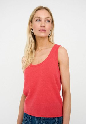 Blonde Frau, die silberne Tropfenohrringe, ein ärmelloses korallenfarbenes Strickoberteil und blaue Denim-Jeans trägt und leicht zur Seite blickt.