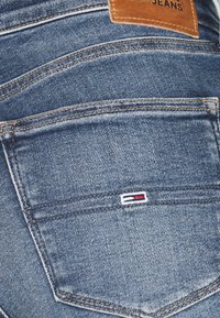 Jeans de mezclilla en azul medio con bolsillos traseros cosidos, que cuentan con un pequeño logo de tela en rojo, blanco y azul cerca del bolsillo derecho.