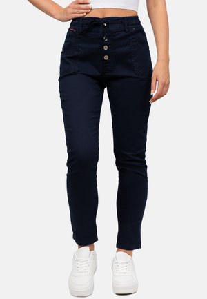 Frau trägt schmale marineblaue Hose mit Knopf und Tunnelzug in der Taille, kombiniert mit weißen Sneakers und einem weißen bauchfreien Top, stehend.
