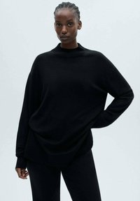 Crni pletena džemper sa visokim izrezom, spuštenim ramenima i opuštenim krojem. Glatka tekstura, u kombinaciji sa odgovarajućim crnim hlačama.