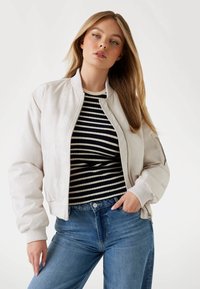 Giacca bomber beige chiaro con colletto e polsini a costine, indossata sopra una maglietta a righe bianche e nere. La modella posa con una mano sui fianchi.