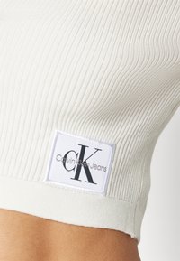 Prążkowany biały crop top z czarną metką logo CK na białym tle, podkreślający dopasowany krój i miękką fakturę.