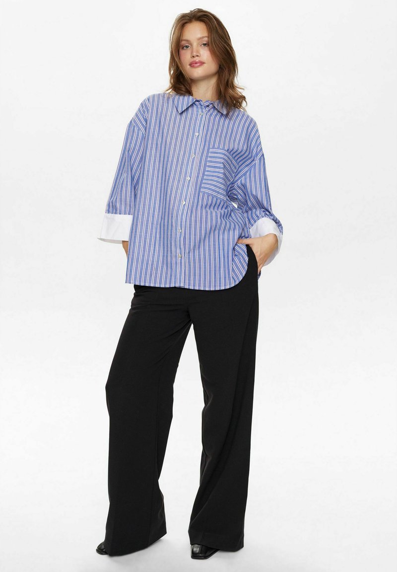 Camicia a righe blu e bianche con una vestibilità rilassata, chiusura a bottoni frontalmente e polsini bianchi arrotolati, abbinata a pantaloni neri a gamba larga.