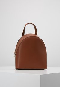 Mochila de cuero marrón con forma redondeada. Cuenta con cierre de cremallera, asa pequeña y textura lisa. Diseño minimalista sin patrones.